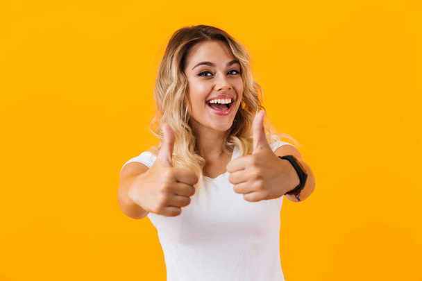 Photo of happy blond woman in basic clothing smiling and showing thumbs up isolated over yellow background - Fotó, kép