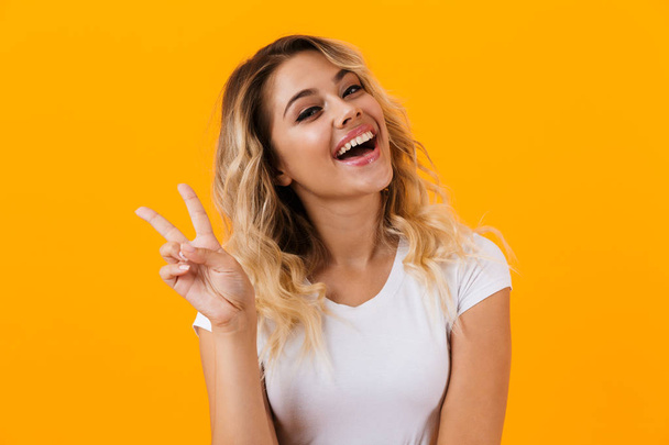 Photo of positive blond woman in basic clothing smiling and showing peace sign isolated over yellow background - Foto, Bild