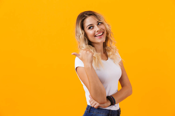 Photo of kind blond woman in basic clothing smiling and pointing finger at copyspace isolated over yellow background - Foto, Bild