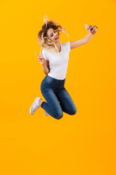 Full length image of young woman jumping and taking selfie photo on mobile phone isolated over yellow background - Фото, зображення