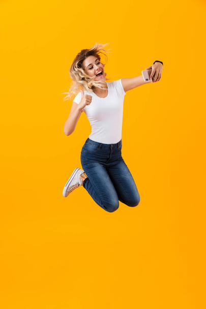 Full length image of beautiful woman jumping and taking selfie photo on mobile phone isolated over yellow background - Фото, зображення