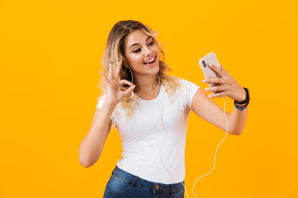 Image of positive woman wearing earphones smiling and taking selfie photo on cell phone isolated over yellow background - Fotoğraf, Görsel