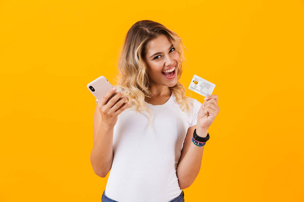 Image of pleased woman holding mobile phone and credit card in hands isolated over yellow background - Valokuva, kuva