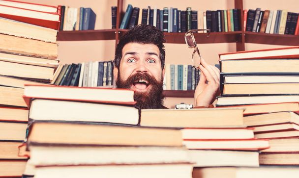 Man on happy face between piles of books in library, bookshelves on background. Scientific research concept. Teacher or student with beard wears eyeglasses, sits at table with books, defocused - Φωτογραφία, εικόνα