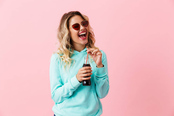 Image of caucasian girl 20s wearing sunglasses drinking soda beverage from glass bottle isolated over pink background - Valokuva, kuva