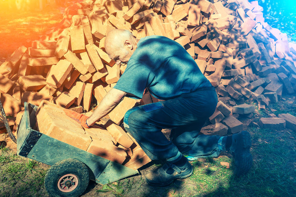 Stary człowiek pracuje na budowie w pobliżu kupę cegieł. Człowiek ładuje cegły na wózku - Zdjęcie, obraz