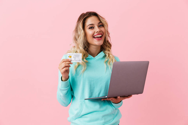 Image of cheerful woman 20s holding silver laptop and credit card isolated over pink background - Fotó, kép