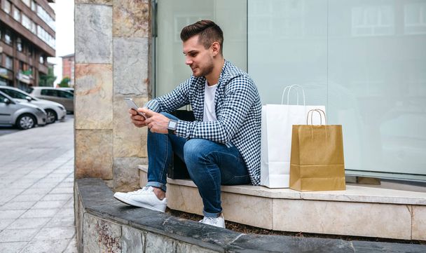 Fiatal ember ül a járda a kirakatba bevásárló táskák - Fotó, kép