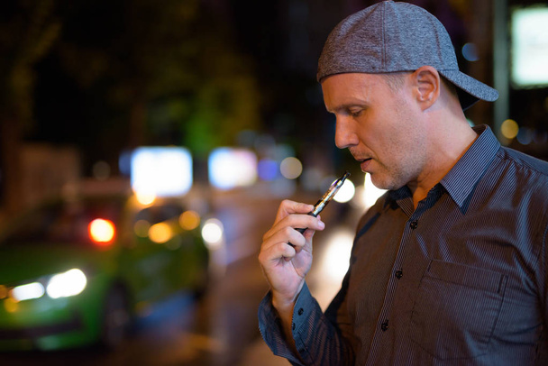 Зрілий чоловік курить електронну сигарету на вулицях вночі
 - Фото, зображення