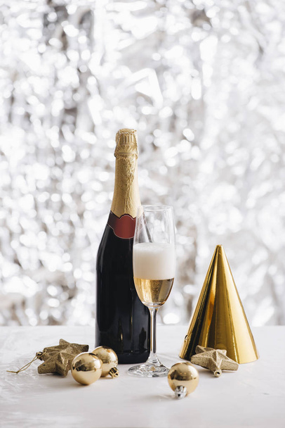 champagne glass and bottle with paper hat and christmas ornaments on silver glitter background - Photo, Image