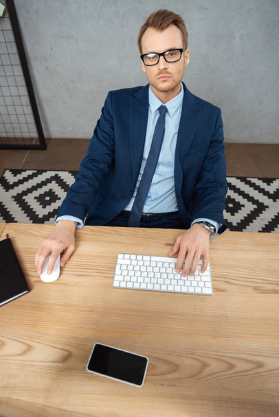 high angle view of businessman in eyeglasses looking at camera and working at table with computer keyboard, mouse and smartphone with blank screen in office  - Foto, Bild