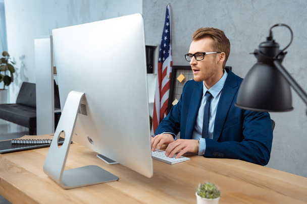 веселый бизнесмен в очках работает за столом с компьютерным монитором в современном офисе
 - Фото, изображение