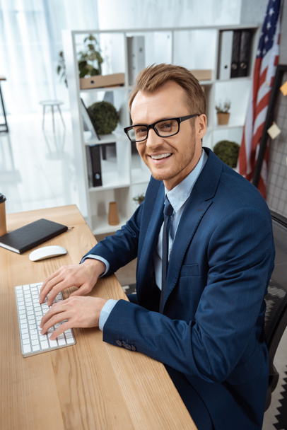 sonriente hombre de negocios en gafas mirando a la cámara y escribiendo en el teclado de la computadora en la mesa en la oficina moderna
  - Foto, imagen
