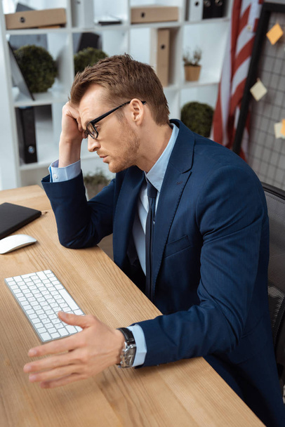jovem empresário cansado em óculos gesticulando à mão à mesa com teclado de computador e mouse no escritório
 - Foto, Imagem