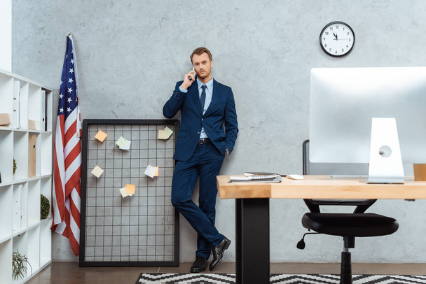 mladý podnikatel v obleku mluví na smartphone poblíž Americká vlajka Wall v moderní kanceláři  - Fotografie, Obrázek
