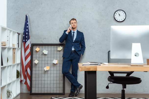 handsome businessman in suit talking on smartphone near american flag by wall in modern office  - Foto, Bild