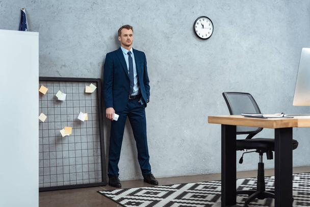confident businessman in suit standing with smartphone near wall in modern office  - Foto, afbeelding