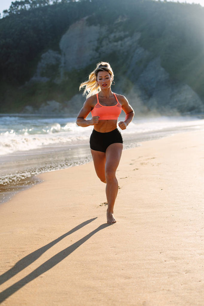 Blonde fitness woman running at the beach in summer morning. - Фото, изображение