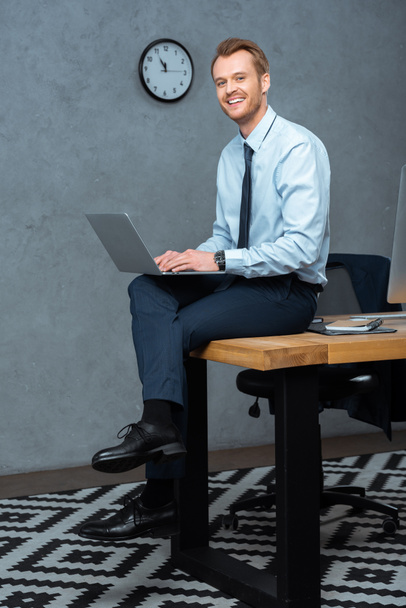uomo d'affari ridente seduto sul tavolo e al lavoro sul computer portatile in ufficio moderno
  - Foto, immagini