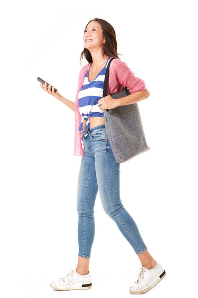Full body side portrait of fashionable young asian woman walking with handbag and mobile phone against isolated white background - Valokuva, kuva