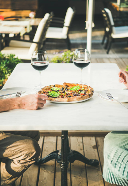 Summer dinner or lunch. Man and woman eating freshly baked Italian vegetarian pizza with vegetables and fresh basil and drinking red wine in outdoor restaurant - Photo, Image