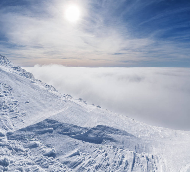 Mountain tops covered with snow above the white heavy clouds. The Sun and blue sky at the background. - Foto, immagini