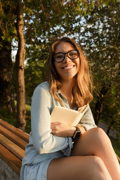Happy young teenage girl with backpack sitting outdoors, holding book - Foto, Imagem