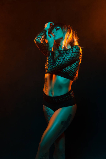 Alternative model with bangs and colored hair poses under teal and orange light wearing a fishnet top - Фото, изображение