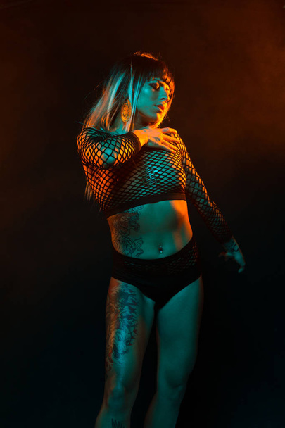 Alternative model with bangs and colored hair poses under teal and orange light wearing a fishnet top - Foto, Imagem