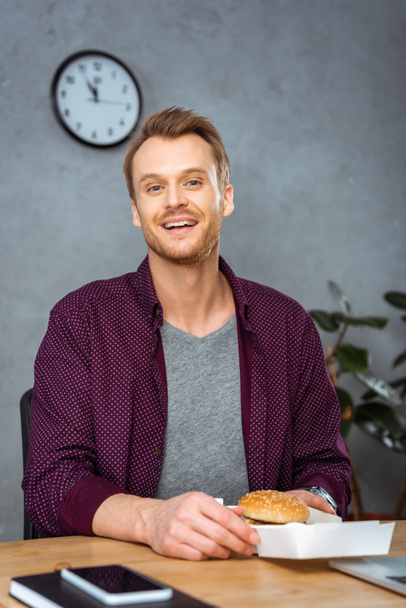 joyful young businessman having lunch with burger at table in modern office  - Fotografie, Obrázek