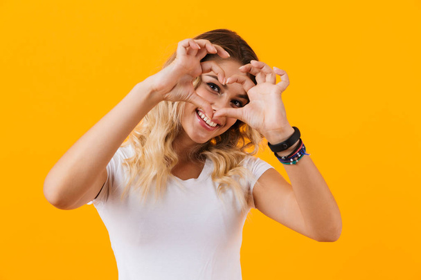 Image of young woman smiling and showing heart shape with fingers isolated over yellow background - Valokuva, kuva