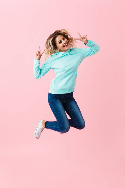 Full length image of happy woman in basic clothing jumping and showing thumbs up isolated over pink background - Foto, imagen