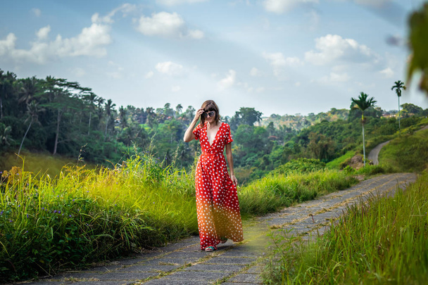 Νεαρή γυναίκα τουρίστας σε ένα lon κόκκινο φόρεμα το περπάτημα στο μονοπάτι τροπικό δάσος. Νησί του Μπαλί. - Φωτογραφία, εικόνα