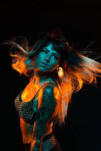 Alternative model with bangs and colored hair poses under teal and orange light wearing a fishnet top - Foto, imagen