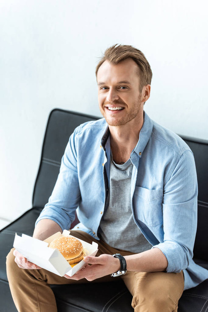 smiling young businessman having lunch with burger while sitting on sofa in office  - Fotó, kép