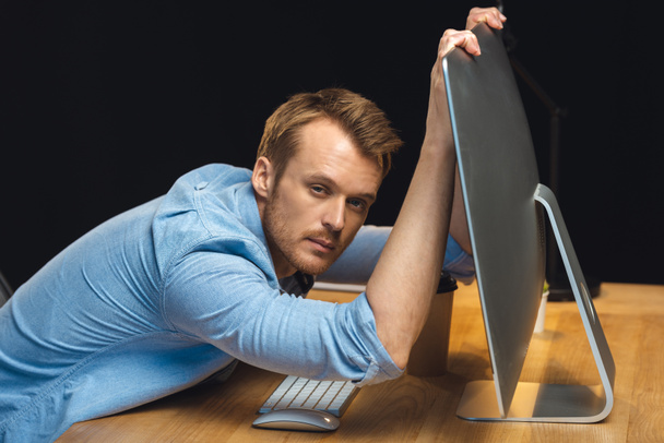 overworked young businessman holding computer monitor and looking at camera during night time in office  - Фото, изображение