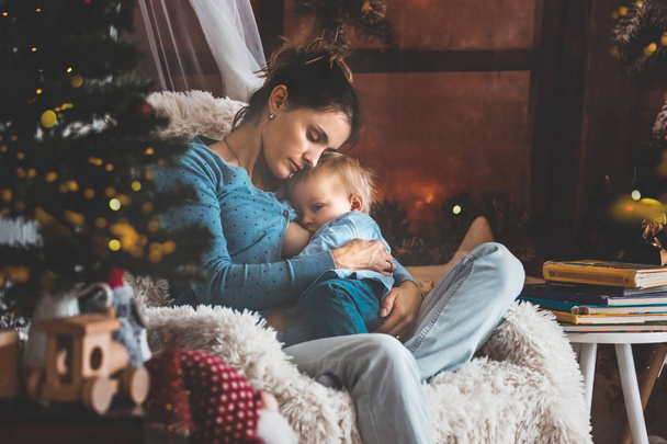 Mother breastfeeding her toddler son sitting in cozy armchair near Christmas tree, wintertime - Zdjęcie, obraz
