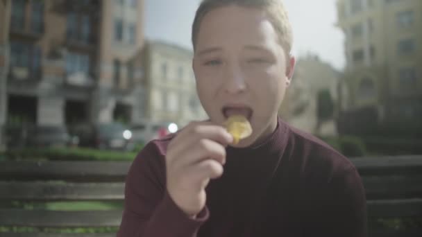 Guy eats chips with pleasure while sitting on a bench - Materiał filmowy, wideo