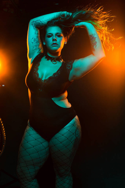 Curvy alternative model with colored hair and lace bodysuit and fishnets poses under blue and orange lighting - Foto, afbeelding