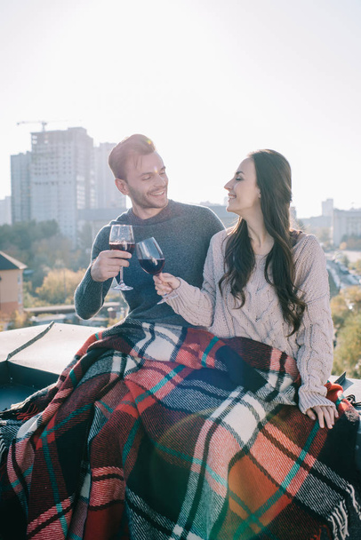 Happy νεαρό ζευγάρι καλύπτοντας σε καρό και τσούγκριζαν ποτήρια κόκκινο κρασί στον τελευταίο όροφο - Φωτογραφία, εικόνα