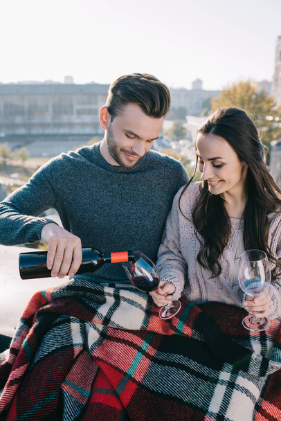 happy young couple covering in plaid on rooftop and pouting red wine into glasses - Foto, Imagen