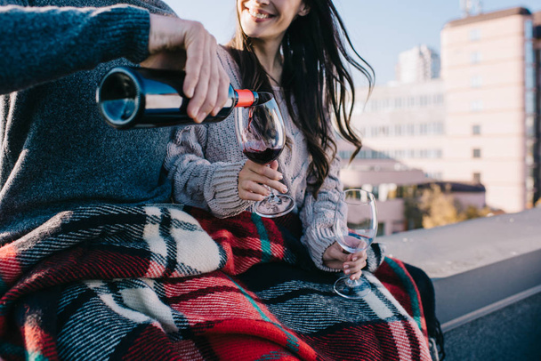 cropped shot of young couple covering in plaid on rooftop and pouting red wine into glasses - Фото, изображение