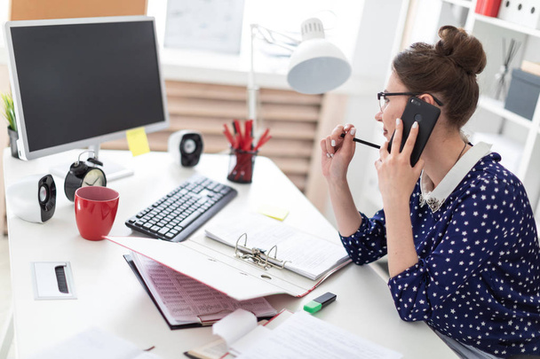 Una joven con gafas se sienta en la oficina en un escritorio de la computadora y está hablando por teléfono. Ante ella en la mesa yacen carpetas con documentos
. - Foto, Imagen