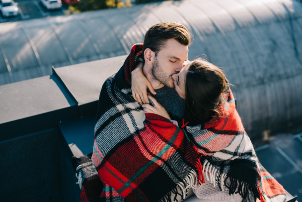 high angle view of beautiful young couple covered in plaid kissing on rooftop - Foto, imagen