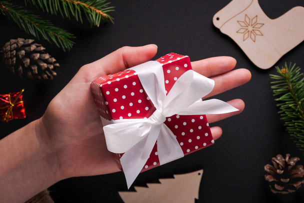 Female hand holding small Christmas gift wrapped in red polka dot paper with white ribbon and bow on black background decorated with spruce twigs, pine cones. Flat lay style - Foto, Imagem