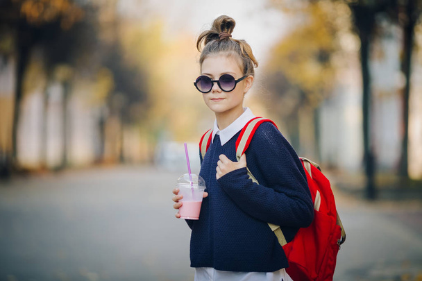 Pretty hipster teen with red bag drinks milkshake from a plastic cup walking street between buildings. Cute girl in sunglasses drinks a drink through a straw. - Foto, imagen