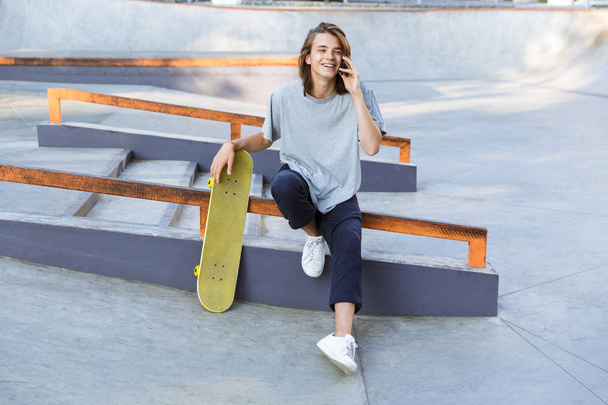Image of handsome young skater guy sit in the park with skateboard talking by mobile phone. - Foto, Bild