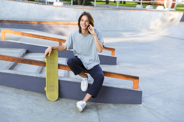 Image of handsome young skater guy sit in the park with skateboard talking by mobile phone. - Foto, immagini