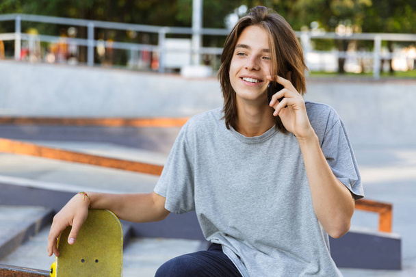 Image of handsome young skater guy sit in the park with skateboard talking by mobile phone. - Valokuva, kuva