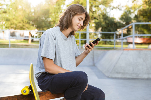 Image of cheerful young skater guy sit in the park with skateboard using mobile phone. - Foto, imagen